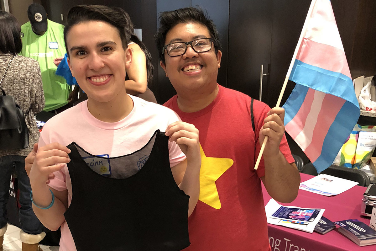 Smiling trans folks hold up a free chest binder and trans pride flag